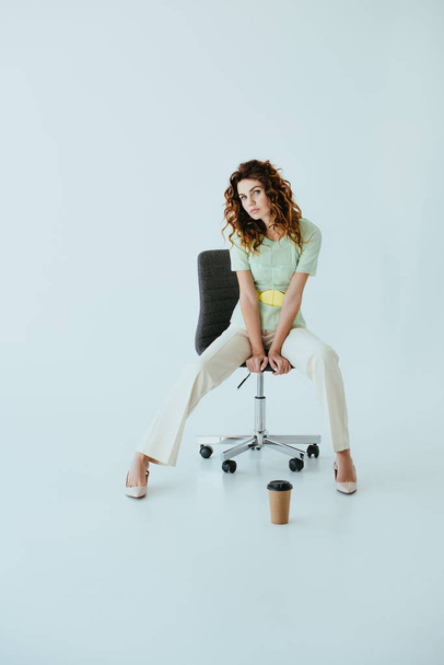 attractive redhead young woman sitting on black office chair near paper cup on grey  - Foto, afbeelding