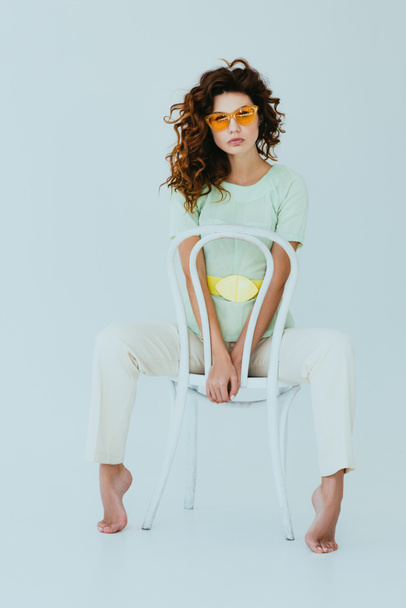 curly redhead young woman in yellow sunglasses sitting on chair and looking at camera on grey  - 写真・画像