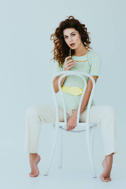 curly redhead young woman holding yellow sunglasses and sitting on chair on grey  - Foto, afbeelding