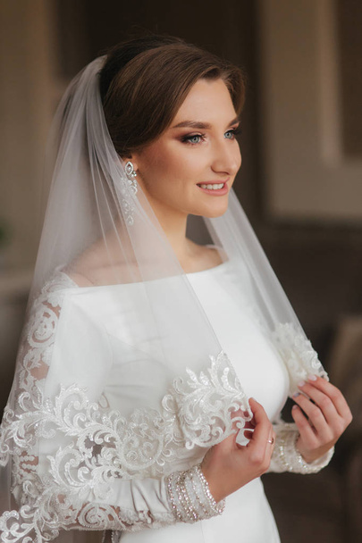 Portrait of beautiful bride standing by the window at home. Charming bride in white wedding dress - Fotografie, Obrázek