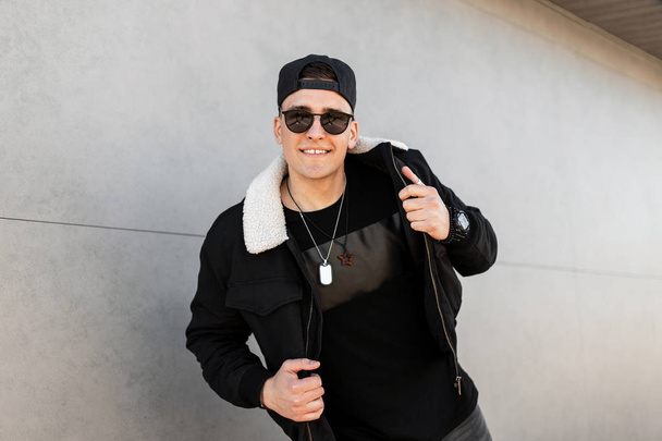 Hombre hipster joven americano feliz en gafas de sol oscuras en chaqueta negra con estilo en una gorra en una camiseta de moda con un amuleto posa y sonríe cerca de un edificio moderno en un día de primavera.Chico alegre de moda
 - Foto, imagen