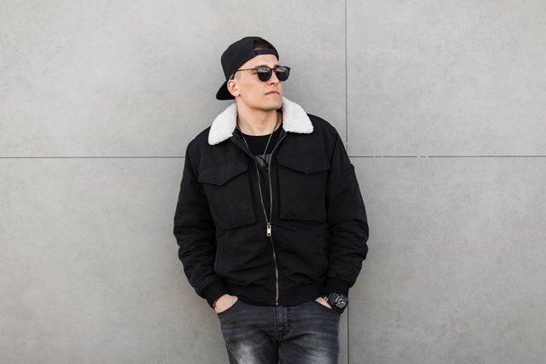 Young attractive hipster man in a black baseball cap in a dark sunglasses in a trendy black jacket with white fur in gray jeans standing near a modern building. Handsome city guy model for a walk. - Photo, Image