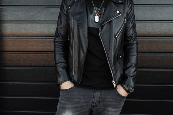 Fashionable young man in a black trendy leather jacket in gray vintage jeans in a stylish black t-shirt posing against a wooden black wall. New collection of men's clothing. Youth style. Close-up. - Φωτογραφία, εικόνα