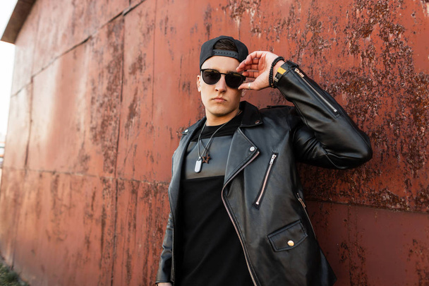 amerikanischer Hipster junger Mann mit dunkler Sonnenbrille in trendiger schwarzer Lederjacke, stylischem Basecap und T-Shirt posiert im Freien in der Nähe der rostigen Metallwand. attraktiver Typ für einen Spaziergang.  - Foto, Bild