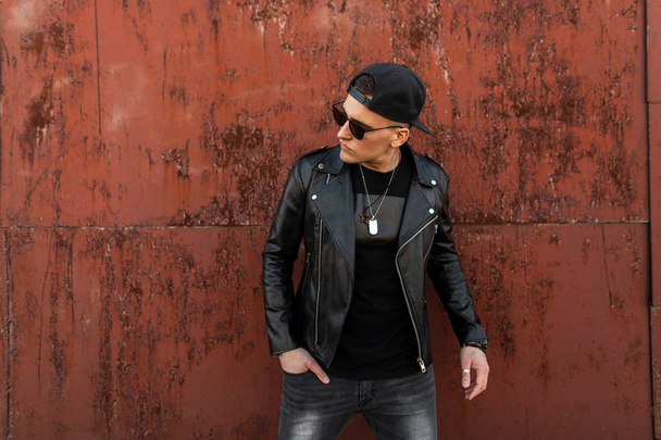 Modern fashionable young hipster man with sunglasses in a stylish leather black jacket in a cap in a vintage T-shirt in gray jeans stands near a metallic red building on a summer day. Handsome guy. - Fotoğraf, Görsel