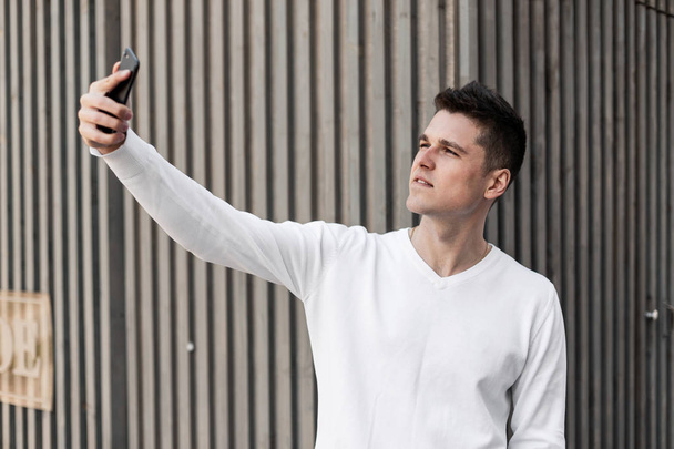 Jonge knappe man met een stijlvol kapsel in een modieus wit shirt maakt selfie staan in de buurt van een grijs houten gebouw van planken in de stad. Een Europese knappe vent. Moderne technologieën. - Foto, afbeelding