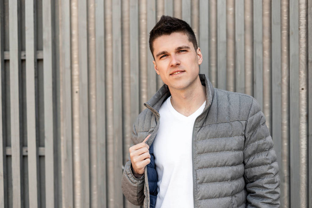 Handsome modern young man with a stylish hairstyle in a fashionable gray jacket in a trendy white T-shirt poses near a vintage wooden wall of boards. Attractive happy guy model for a walk. - Fotó, kép