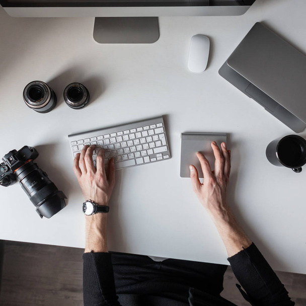Young successful photographer is sitting at the table and typing on the keyboard. Top view of the desktop on which there is a keyboard, camera, laptop and professional lenses to the camera.  - Foto, immagini