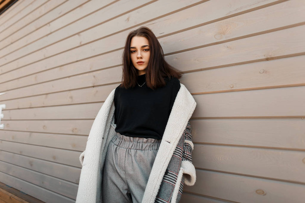 European fashionable young woman model in gray stylish pants in a black shirt in an checkered jacket posing standing near a wooden building. Cute girl outdoors. Spring collection of women's clothing. - 写真・画像