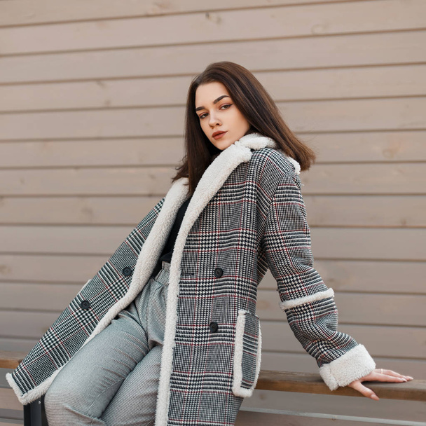 Pretty sexy young woman in a elegant checkered jacket in a shirt in gray vintage pants sits and relaxes on a spring day near a vintage wooden wall outdoors. Sensual modern girl model rest. - Foto, afbeelding