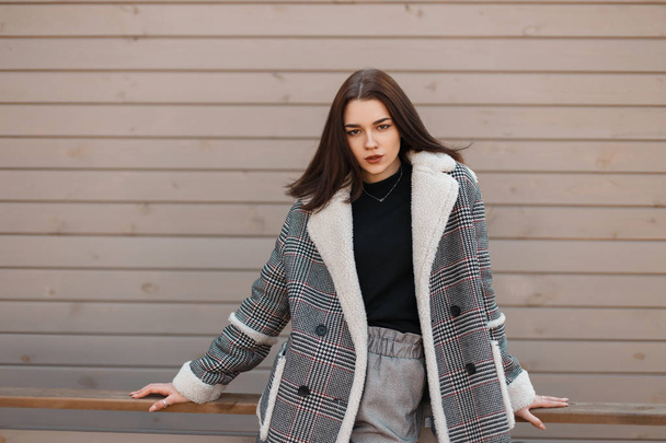 Beautiful modern young woman in trendy black T-shirt in a gray elegant checked jacket in fashionable vintage pants is standing outdoors near the wooden wall. European girl fashion model. Youth. - Foto, afbeelding