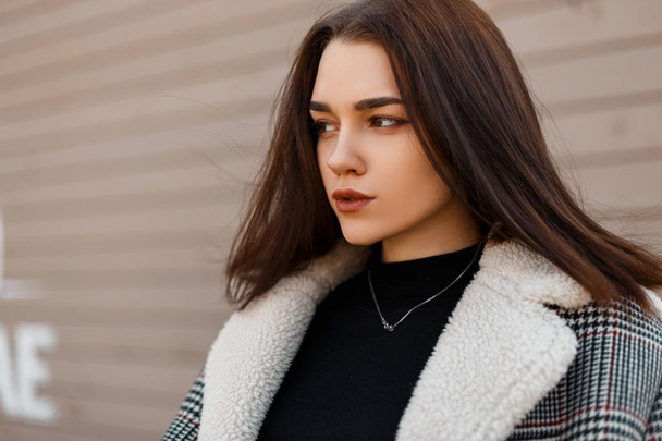 Portrait of a cute beautiful young brunette woman with natural make-up in a fashionable checkered jacket with fur in a stylish black t-shirt near a wooden beige wall outdoors spring day. Pretty girl. - Photo, Image