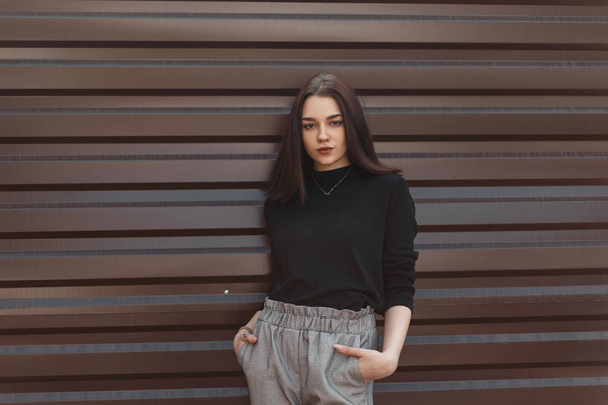 Young pretty beautiful woman in black vintage sweater in fashionable gray checkered pants poses standing near a metallic brown fence in the city. Attractive modern girl model outdoors. - Photo, Image