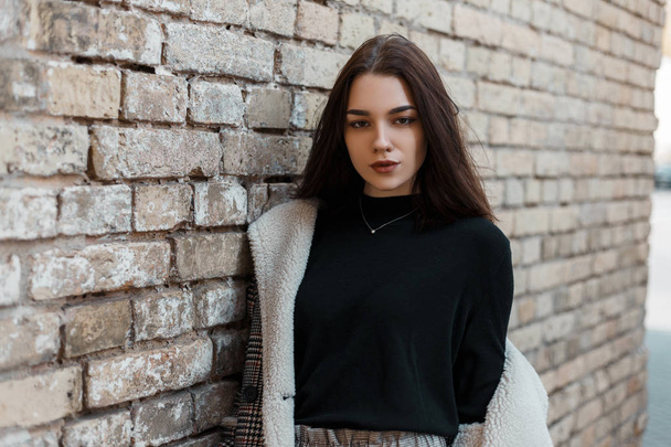 Pretty young brunette woman with a natural make-up in a fashionable plaid jacket with white fur in a black T-shirt is resting standing near a brick vintage house in the city. Stylish beautiful girl. - Foto, afbeelding