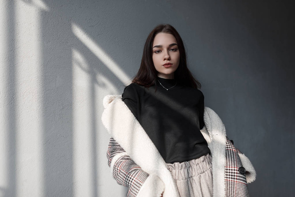 Fashionable pretty young woman in a stylish gray checkered jacket in vintage pants in a black stylish shirt is standing indoors near the wall. Attractive brunette girl is resting in the sunny room. - Zdjęcie, obraz