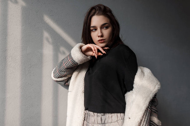 Elegant modern attractive brunette woman in a checkered vintage coat in a stylish sweater in gray pants is standing in a room with concrete walls. Pretty beautiful girl model relaxes in the sun. - Foto, imagen