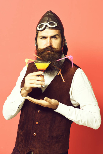 handsome bearded pilot or aviator man with long beard and mustache on serious face holding glass of alcoholic beverage in vintage suede leather waistcoat with hat and glasses on red studio background - Zdjęcie, obraz