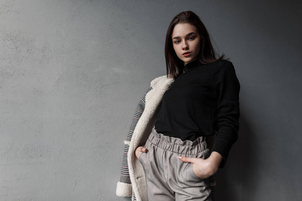 Ευρωπαϊκό μοντέλο μόδας μιας νεαρής γυναίκας με γκρίζο κομψό παντελόνι σε ένα μαύρο πουκάμισο σε ένα καρό μπουφάν που ποζάρει στέκεται κοντά σε ένα vintage τσιμεντένιο τοίχο. Χαριτωμένο κορίτσι. Ανοιξιάτικη συλλογή ενδυμάτων γυναικών. - Φωτογραφία, εικόνα