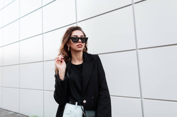 European pretty young hipster woman in trendy sunglasses in a vintage coat in white jeans with a bag stands near a gray building in the city. Beautiful girl for a walk. Stylish women's clothing. - Photo, Image
