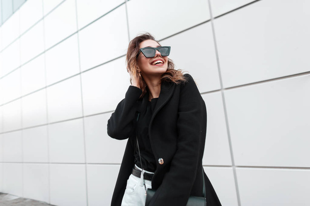 Vrolijke jonge vrouw hipster in zonnebrillen in een trendy jas in een T-shirt met een stijlvolle lederen handtas met een schattige glimlach in Vintage jeans staat in de buurt van een modern grijs gebouw in de stad. Gelukkig meisje - Foto, afbeelding