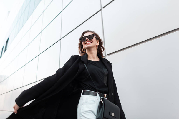 Giovane donna hipster felice in cappotto nero lungo alla moda in jeans bianchi in eleganti occhiali da sole con una borsa di pelle viaggia per la città vicino al muro moderno. Ragazza positiva che viaggia lungo la strada
. - Foto, immagini