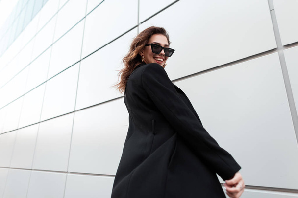 Örömteli fiatal csípő nő a divatos, hosszú, fekete kabát sötét stílusos napszemüveg, pozitív mosollyal utazik a város körül, közel a modern fal. Boldog lány sétál az utcán. - Fotó, kép