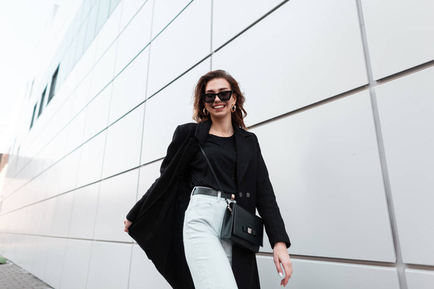 Joven mujer hipster con estilo positivo en gafas de sol en un abrigo elegante en una camiseta en jeans vintage con un bolso de cuero está caminando y sonriendo cerca de un edificio moderno en la ciudad.Chica alegre camina
 - Foto, Imagen