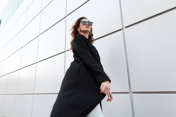 Jonge mooie hippe vrouw in een vintage lange zwarte jas in modieuze zonnebril gaat op een warme lentedag in de buurt van een modern gebouw. Stijlvol aantrekkelijk meisje reist door de stad. Retrostijl. - Foto, afbeelding
