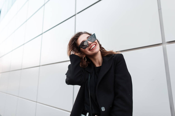 Young funny beautiful hipster woman in sunglasses in a vintage black coat in a T-shirt with a positive smile poses and laughs while standing near a modern gray building in the city. Joyful girl. - Фото, изображение