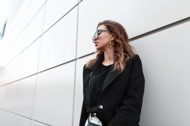 European urban young women hipster in fashionable black coat in a t-shirt in dark trendy sunglasses in jeans with a bag resting standing near the modern wall outdoors. Stylish girl. Retro style. - Фото, зображення