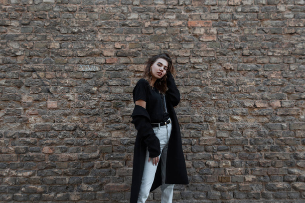 Europese stijlvolle jonge vrouw in een vintage zwarte lange jas in witte jeans in een modieuze t-shirt poseren staande in de buurt van de bakstenen bruine muur buitenshuis. Mooi aantrekkelijk meisje voor een wandeling. Women's Fashion - Foto, afbeelding