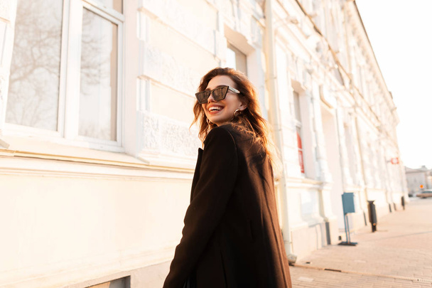 Young cheerful hipster woman with a positive smile in black sunglasses in an elegant coat posing near a white vintage building in the city on a bright sunny day. Joyful girl model enjoys the sunset. - Valokuva, kuva