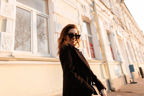 Pretty cheerful hipster young woman in dark stylish sunglasses in long fashionable coat with a handbag posing on the street near a vintage building. Joyful attractive girl model travels at sunset. - Foto, Imagem