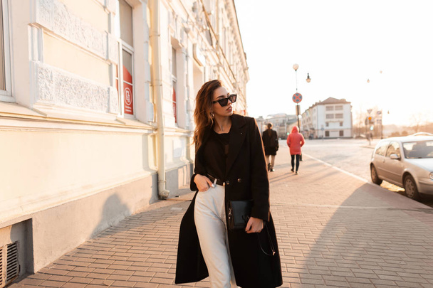 European pretty young hipster woman in a vintage black long coat in white jeans in a t-shirt posing standing in the city near the white buildings of the wall. Stylish girl model enjoys the sunshine.  - Foto, Imagem
