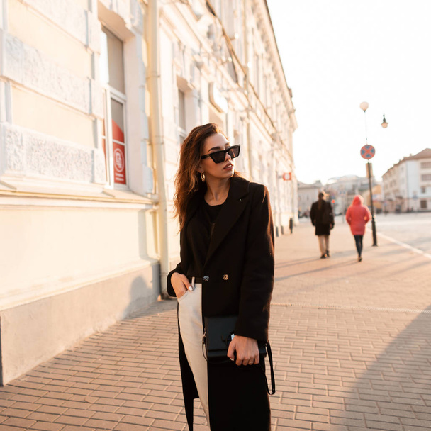 Modern beautiful young woman hipster in an elegant coat in a T-shirt in white jeans with a leather bag in sunglasses posing near a vintage building outdoors. Attractive girl enjoys sunny day. Youth. - Fotoğraf, Görsel