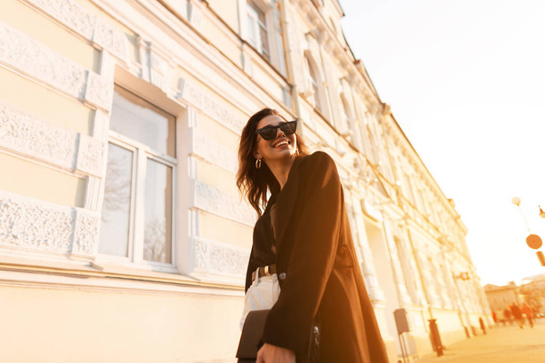 Cheerful young hipster woman in trendy clothes in stylish sunglasses walks around the city and enjoys the bright orange spring sunset. Beautiful joyful girl travel on a sunny day outdoor. Modern style - Valokuva, kuva