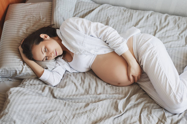 若い妊娠中の女性はベッドの上に横たわっています彼女のおなかを抱きます,健康的な妊娠 - 写真・画像