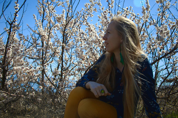 Una chica con el pelo largo rubio y los ojos verdes sonríe mirando a la izquierda y tocándose el pelo delante de un árbol en flor
. - Foto, imagen