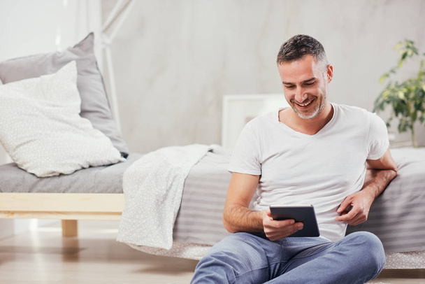 Handsome Caucasian middle-aged man sitting on the floor in bedroom and leaning on bed. In hands tablet. - Photo, Image