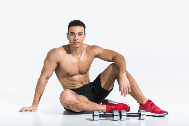 handsome sportive mixed race man sitting on floor near dumbbells on white - Foto, immagini