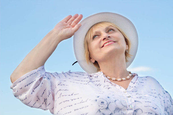 Ευτυχισμένο χαμογελάει συνταξιούχος γυναίκα ρολόγια ουρανό κρατώντας το λευκό καπέλο της - Φωτογραφία, εικόνα