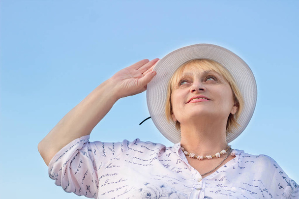 Χαρούμενος χαμογελαστός ηλικιωμένη γυναίκα ρολόγια ουρανό κρατώντας το λευκό καπέλο της - Φωτογραφία, εικόνα