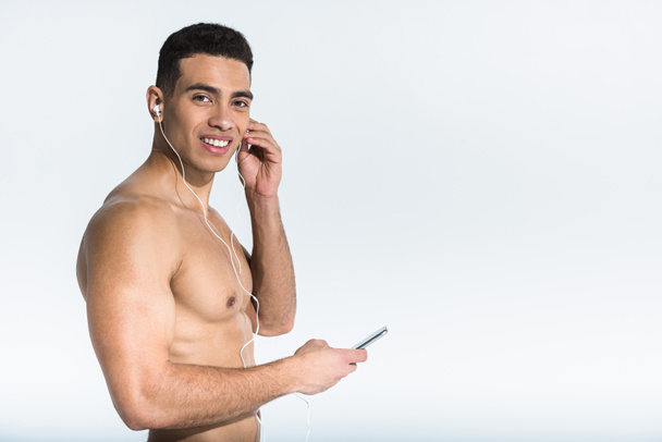 handsome athletic mixed race man listening music in earphones and looking at camera on white - Foto, afbeelding