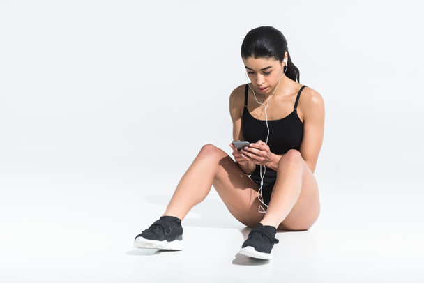 pretty african american girl in sportswear sitting on floor and listening music in earphones on white - Photo, Image