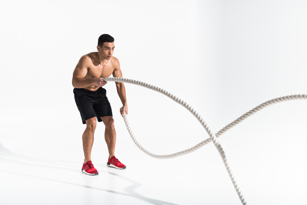 handsome mixed race man in black shorts and red sneakers doing exercise with ropes on white - 写真・画像