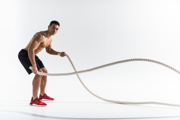 handsome mixed race man in black shorts and red sneakers training with ropes on white - Photo, Image