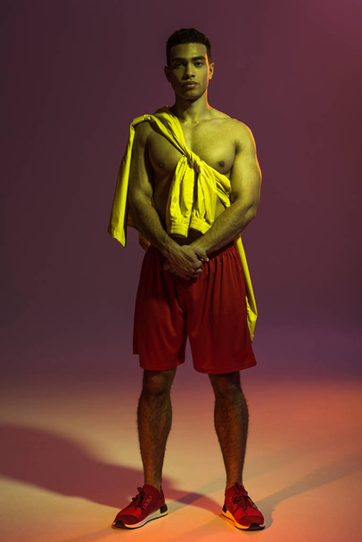 apuesto hombre de raza mixta en pantalones cortos rojos, y chaqueta amarilla atada en el torso mirando a la cámara en el fondo oscuro
 - Foto, Imagen