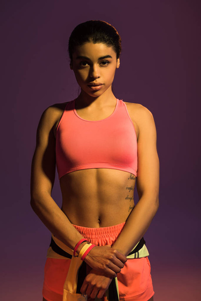 pretty african american girl in pink sport bra looking at camera on purple background - Photo, Image