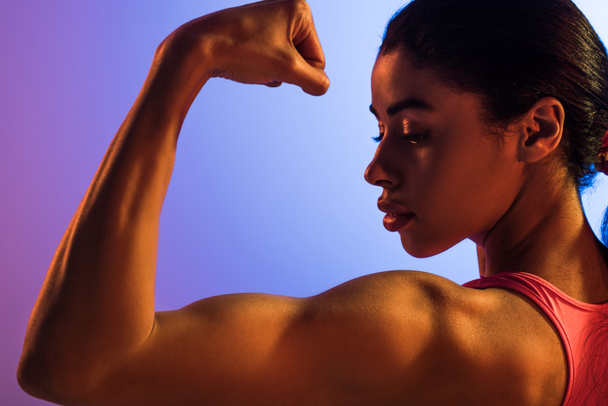 beautiful athletic african american girl demonstrating biceps on purple and blue gradient background - 写真・画像