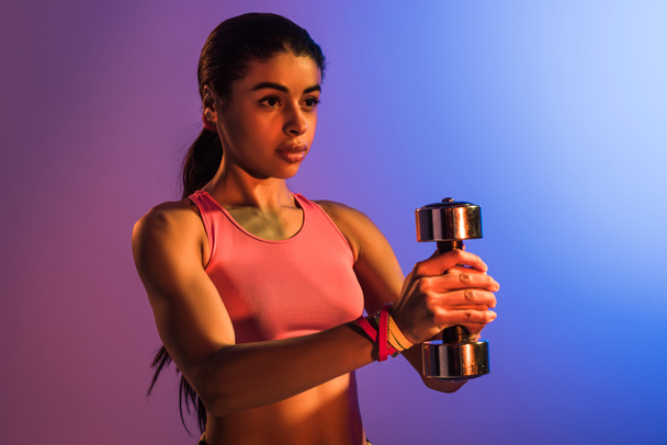 attractive african american woman in pink sports bra holding dumbbell on purple and blue gradient background - Fotó, kép